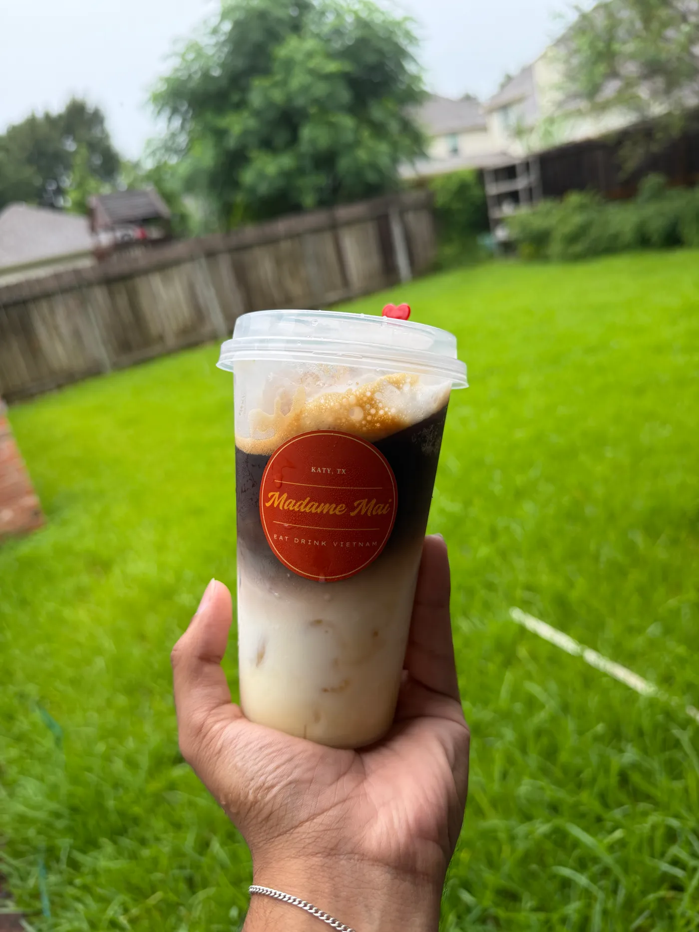 Hand holding a layered iced Vietnamese coffee in a Madame Mai cup over a green lawn